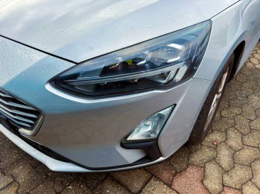 Vücudunda su damlacıkları olan gümüş bir araba tuğla bir yüzeye park edilmiştir. Odak noktası ön far ve hafif yağmur sırasında arabanın dış tasarımı..