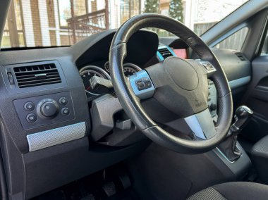 Bir kişi direksiyona ve gösterge paneline bakarak bir arabanın sürücü koltuğunda oturur. Araba, pencereden içeri güneş ışığı giren bir yere park edilmiş..