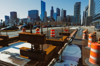 Chicago şehir merkezinde yol yapımı devam ediyor. İşçiler, trafik konileri ve aletler işlek bir caddede mevcuttur. Güneşli bir kış gününde şehrin silueti arkalarındadır..