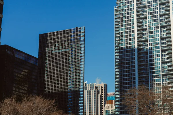 Açık mavi bir gökyüzünün altında iki yüksek bina yan yana duruyor. Bir binanın camları var. Duman uzaktaki bir binadan geliyor..