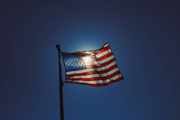 A flag is waving at dusk against a clear blue sky. The stars and stripes are visible as the flag moves in the wind, standing out against the darkening background.