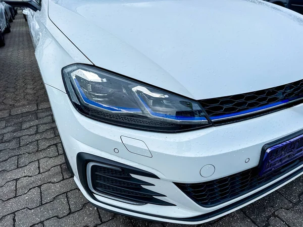 Beyaz bir arabanın önü bir araç parkında park edilmiş. Zemin ıslak ve hava bulutlu. Arabanın parlak farları olan parlak bir tasarımı var..