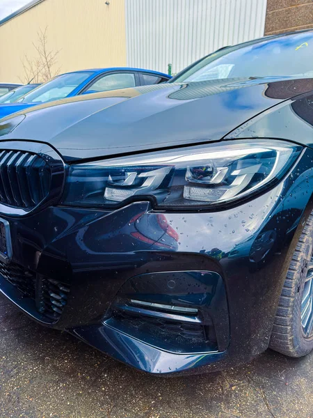 Birçok arabanın arasında siyah bir araba park edilir. Yağmur damlaları yüzeyinde görülebilir. Bulutlu bir gün ve çevrede metal binalar var..