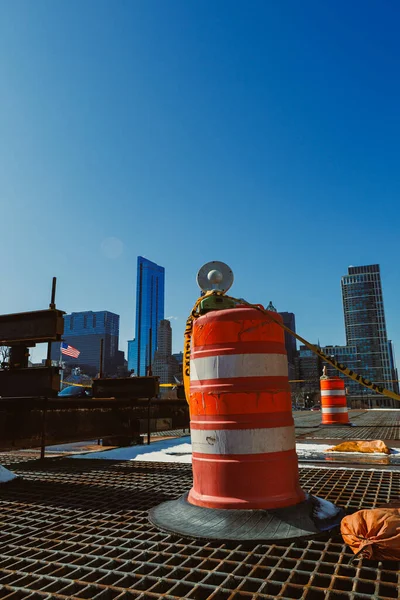 Bir inşaat alanı trafik konileri ve turuncu fıçılarla gösteriliyor. Şehrin ufuk çizgisi arka planda gün boyunca parlak bir gökyüzünün altında görünür..
