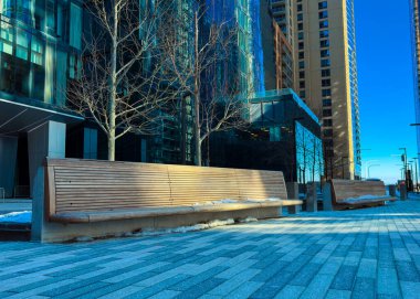 Aydınlık bir kış günü, yüksek binaların arasında ağaçların altındaki ahşap bankların yanındaki patikada yürüyen bireyler görülüyor..