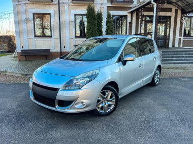 Gümüş renkli bir hatchback araba bir binanın dışında pürüzsüz bir yüzeye park edilmiştir. Araba, parlak vücudu güneş ışığını yansıtırken öne bakıyor. Yakınlarda yeşil ağaçlar görülebilir..