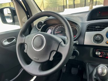 Bir arabanın ön koltuğunda bir direksiyon ve torpido gözlenir. Dışarıda pencerelerden kar görünüyor, bu da kış koşullarını gösteriyor. Araba park halinde..