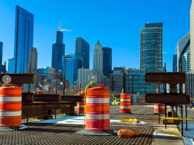 İnşaat bariyerleri şehrin ufuk çizgisinin önündeki bir alanı kapatıyor. Mavi bir gökyüzünün altında arka planda uzun binalar duruyor. Sahne devam eden kentsel gelişimi gösteriyor.