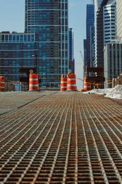 İşçiler şehir bölgesinde bir bina inşa ediyorlar. Çelik kirişler ve beton mevcut ve inşaat alanında turuncu bariyerler var. Sitenin etrafındaki yüksek binalar.