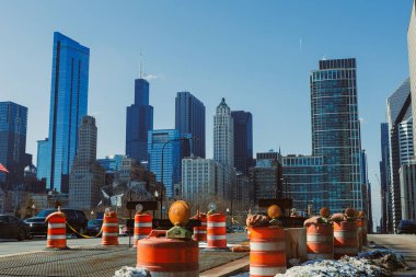 İşçiler turuncu trafik konileriyle dolu işlek bir şehir caddesinde inşaat işindeler. Uzun gökdelenler arka planda yükseliyor Chicago şehir merkezinde açık mavi bir gökyüzünün altında.