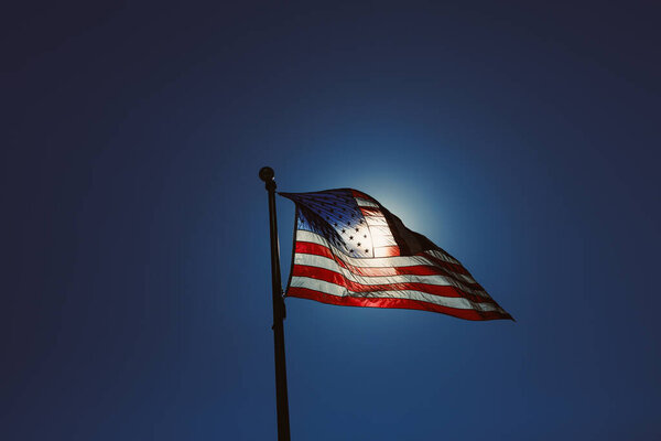 A flag of the United States stands tall against the blue sky. The sun shines bright behind the flag, creating a vivid display. The colors of the flag are visible in the light.