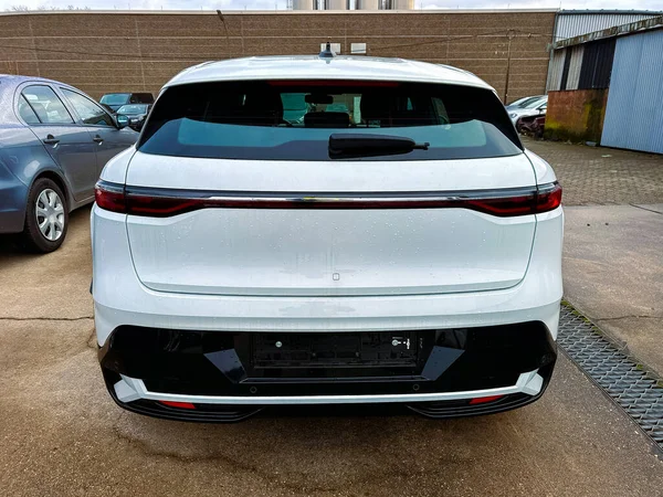 Modern bir araba çok park edilir. Aracın arkası parlak bir tasarımı gösteriyor. Yakınlarda başka arabalar da görülebilir. Gökyüzü bulutlu. Arazide farklı yüzey ve binalar var..