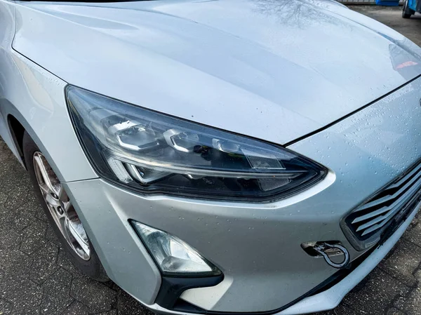 Dışarıda park edilmiş gümüş bir araba var. Odak noktası ön taraf, farlar ve kaput dahil, yüzeyinde yağmur damlaları var. Hava yağmurlu..