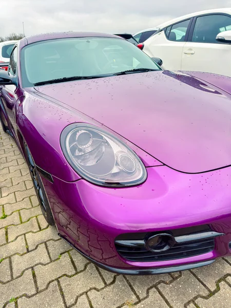 Bulutlu bir günde mor bir spor araba diğer araçların arasına park edilir. Yüzeyindeki yağmur damlaları donuk gökyüzünü yansıtır. Arabanın eşsiz bir tasarımı var ve göze çarpıyor..