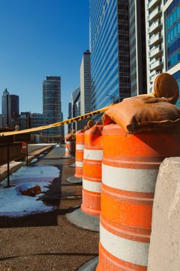 Trafik engelleri mavi bir gökyüzünün altında bir şehir sokağının kenarına kurulur. Bariyerlerin arkasında yüksek binalar yükseliyor. Kar yakınlarda görüldü..