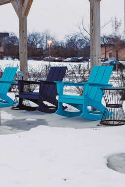 Üç farklı renkte sandalye bir verandaya kurulur ve etrafları karla çevrilidir. Bölge kış belirtileri gösteriyor ve güneş batmaya başlıyor. Çevrede seyrek çalılıklar ve park edilmiş birkaç araba var..