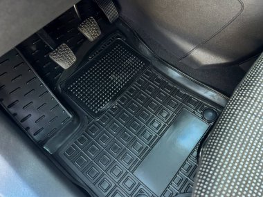 Arabanın zemininde lastik paspas, gaz ve fren pedalları ve koltuğun bir kısmı görünür. Güneş ışığı iç kurulumun ayrıntılarını vurgular.