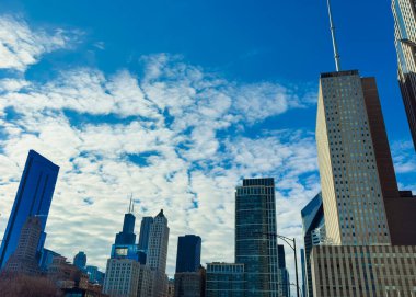 Şehrin silueti parlak mavi gökyüzüne uzanan yüksek binaları gösteriyor. Kabarık bulutlar gökyüzünü doldurarak cam ve çelik yapılarla bir kontrast oluşturuyor. Şehir yaşamının manzarası aşağılarda sergileniyor..