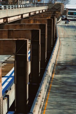 Bir otoyol üst geçidi, onu destekleyen büyük metal sütunların olduğu yüksek bir manzaradan görülebilir. Arabalar aşağıda yol boyunca ilerliyor ve otoyolun yanında bir nehir akıyor..