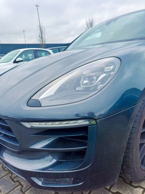 Koyu mavi bir arabanın ön manzarası farları ve ızgarayı gösteriyor. Diğer arabalar gün boyunca bulutlu bir gökyüzünün altında arka planda park halindedir..