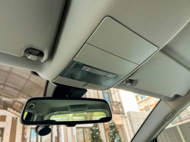 Tepedeki konsol ve dikiz aynası park halindeki bir arabanın içinde görünür. Işık arabaya giriyor, yerleşim alanının bir kısmını bazı binalarla birlikte gösteriyor..