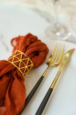 Altın bir yüzükle sarılmış bir bez peçete beyaz bir tabakta çatal ve bıçağın yanında yatıyor. Sahne, basit detaylara odaklanarak yemeğe hazır bir yemek düzenini gösteriyor..