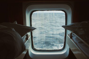 Kabin, dışarıda su olan bir pencere gösteriyor. Gökyüzü bulutlu görünüyor ve içinde yastıklı iki yatak var. Su damlacıkları camda..