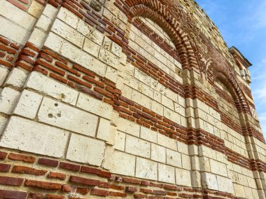 Yapı, kırmızı tuğlalar ve beyaz taşlardan yapılmış bir duvarın detaylı bir görüntüsünü gösteriyor. Duvarda kemerli açıklıklar ve yaş belirtileri var. Gökyüzü açık ve mavi.