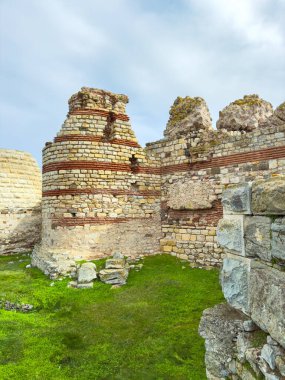 İnsanlar taştan yapılmış harabelerin etrafında yürüyor, kırık duvarları ve çimenli zemini inceliyorlar. Güneş ışığı eski yapıların üzerinde parlıyor, yaşlarını gösteriyor..