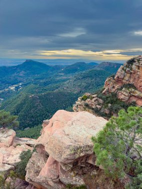 Yüksek açı kayalık araziyi ve uzun ağaçları gösterir. Gün batımında bulutlu bir gökyüzünün altında uzanan bir manzara..