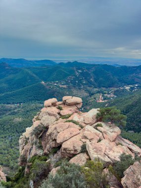 Yeşil tepelere ve vadilere tepeden bakan kayalık bir dağın tepesinde bir kişi duruyor. Gökyüzü gri tonlarla bulutlu ve manzara uzak ağaçları ve küçük yerleşim yerlerini gösteriyor..