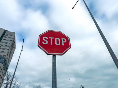 Kırmızı bir dur işareti yüksek binaların ve sokak lambalarının yanındaki uzun bir direğe konur. Gökyüzü bulutlu ve şehir merkezinde gündüz vakti görünür bulutlar var..