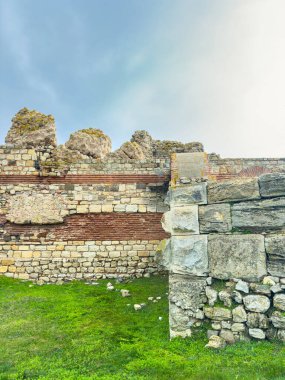 Tarihi kalıntıların bir parçası olan taş bir duvar. Duvarın önünde çimen parçaları ve yukarıda mavi bir gökyüzü var..