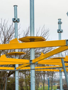 İşçiler parkta sarı bileşenleri olan bir oyun alanı inşa ediyorlar. Metal kutuplar yerden yükseliyor ve bazı parçalar hala bir araya getiriliyor. Bulutlu bir gün..