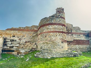 Kırık taşlar etrafa saçılmış eski kalıntılar çimenli bir alanda duruyor. Yapı, mavi gökyüzünün altındaki duvarların ve dairesel bölümlerin parçalarını gösteriyor. Bu site tarihi ve zamanı yansıtır.