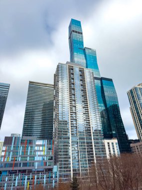 Şehir manzarası üzerinde birkaç yüksek bina yükselir. Bulutlu bir günde gökyüzünü ve etrafındaki yapıları yansıtan cam yüzeylere sahiptirler. İnsanlar aşağıda hareket eder..