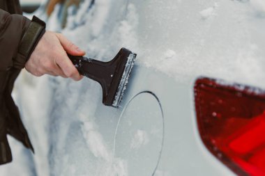 Karlı bir ortamda bir insan arabanın arkasından karı kazır. Kar yüzeyinde birikerken araba dışarıda park halinde..