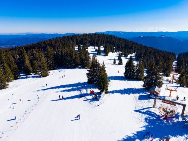 İnsanlar ağaçlar ve karla çevrili bir dağ otelinde yamaçlardan kayak yapıyorlar. Güneş parlıyor ve gökyüzü açık..