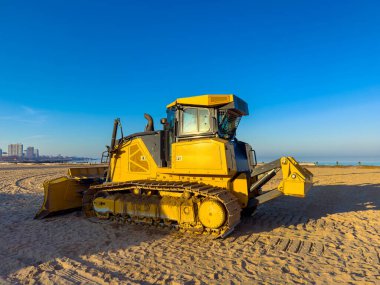 Büyük sarı bir buldozer kumları hareket ettirmek ve yüzeyi insanlar için düzleştirmek için sahilde çalışır. Okyanus arka planda görünür ve gökyüzü berrak..