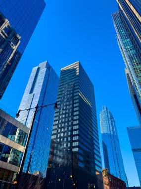 Cam yüzeylerde yansımaları olan bir şehirde gökyüzüne doğru yükselen uzun binalar. Olay yeri, gün ortasında canlı bir şehir alanını gösteriyor..