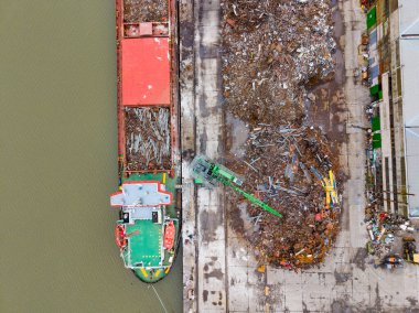İşçiler yakındaki hurdalıkta çeşitli metal parçalarını yönetirken bir kargo gemisi rıhtımda duruyor. Bölge, gün boyunca sanayi faaliyetleri gösteriyor..