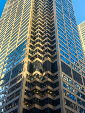 Bir şehirde yüksek bir bina ayakta duruyor. Dış yüzeyin görsel bir etki yaratan bir şablonu var. Cam pencerelerde gökyüzünün yansımaları beliriyor. Dışarısı aydınlık..