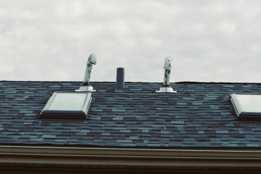 İki çatı penceresi, havalandırma için iki boruyla birlikte karanlık bir çatıda duruyor. Yukarıdaki gökyüzü çoğunlukla bulutlu ve gri. Olay yeri bir yerleşim alanını gösteriyor..