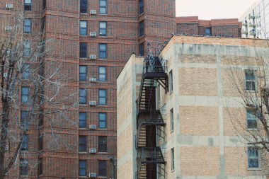 İnsanlar yan yana iki apartman görüyor. Birinin yangın çıkışı ve bazı pencerelerde klima var. Yapraksız ağaçlar ön planda görünür..