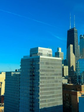 Yüksek binalar Chicago 'nun yüksek bir noktasından manzarayı dolduruyor. Açık mavi gökyüzü, şehir hayatı devam ederken parlak bir manzara yaratıyor. Mimari yapılar birbirine yakın duruyor..