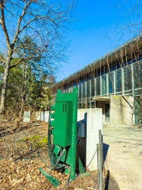 Büyük cam pencereli modern bir binanın yanında yeşil bir hizmet kutusu bulunmaktadır. Kuru yapraklar yere saçıldı. Ağaçlar çıplak, gökyüzü açık ve mavi.