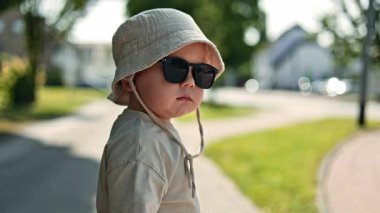 Şık bir güneş şapkası ve sevimli bir güneş gözlüğü takan küçük sevimli bir bebek güzel, güneşli bir günde, neşeli bir oyuncak arabasıyla canlı dışarıyı keşfediyor.