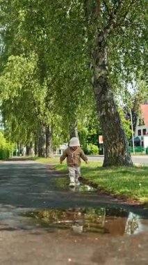 Lastik çizmeli tatlı 2 yaşındaki çocuk yağmurdan sonra su birikintilerinde koşuyor. Küçük çocuk su birikintisinde duruyor ve oynamaktan hoşlanıyor. Çocuk sokakta su birikintisine atlıyor..