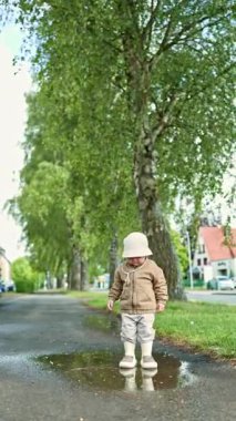 Lastik çizmeli tatlı 2 yaşındaki çocuk yağmurdan sonra su birikintilerinde koşuyor. Küçük çocuk su birikintisinde duruyor ve oynamaktan hoşlanıyor. Çocuk sokakta su birikintisine atlıyor..