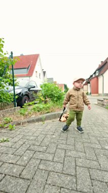 Şapkalı ve ceketli bir çocuk sokakta arabayla yürüyor. Çocuk plastik bir arabayla oynuyor. Tasmalı çocuk arabası. Erkekler için oyuncaklar. Yürüyüşe çıkmış 2 yaşında şık bir çocuk..